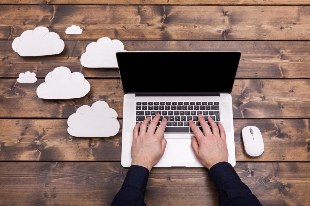 Hände an einem Laptop auf Holztisch mit stilisierten Papierwolken als Symbol für Cloud-Technologien